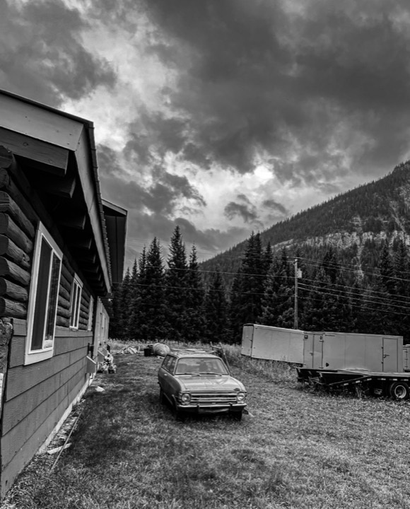 Black-and-white photo of a log cabin beside an old car and a semi-trailer, with storm clouds over forested mountains